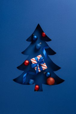 Dikey mavi kağıttan bir Noel ağacı kesimi renkli süsler ve çizgili bir hediye içerir, tebrik kartları ve mevsimsel promosyonlar için uygun bir bayram motifi yaratır