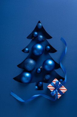 Dikey mavi Noel ağacı kesimi, parlak süsler ve çizgili bir hediye, mavi bir zemin üzerinde kıvrımlı kurdeleyle vurgulanmış, yeni yıl için bayram havasını ve Noel selamlarını iletiyor.