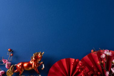 Altın bir at heykeli, kırmızı katlanan yelpaze ve pembe çiçeklerin olduğu canlı dekoratif bir sahne. Derin mavi arka planda, Çin Yeni Yılı kutlamalarının somutlaştırılması ve sıcak karşılama duygusuyla.