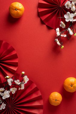 Canlı, dikey Çin Yeni Yılı sahnesinde parlak kırmızı hayranlar, mandalinalar ve zarif beyaz çiçekler koyu kırmızı arka planda sergilenir.