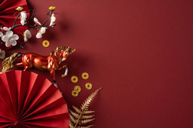 Kırmızı bir yelpazenin, altın at heykelciğinin, çiçeklerin ve bozuk para aksanının yer aldığı yeni yıl şenlikli bir Çin sahnesi şans ve kutlama amaçlı zengin bir kırmızı zemin üzerine kuruludur.