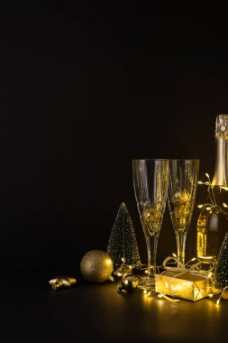 Karanlık dikey arka plan, şampanya bardakları, bir şişe, yanıp sönen ışıklar, küçük hediyeler ve sıcak, şenlikli bir ortam yaratan dekoratif ağaçlardan oluşan altın bir tatil tablosunu ön plana çıkarıyor.