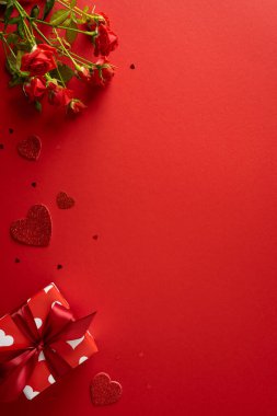 Kırmızı dikey arka planda bir buket gül, dağınık parıltılı kalpler ve kurdeleli bir hediye kutusu bulunuyor. Sevgililer Günü havası ve romantizmi yansıtıyor.