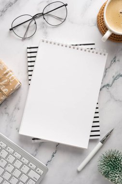 Boş bir defter, yuvarlak bardaklar, bir klavye, bir kalem, hediye paketi ve Noel ve yeni yıl hazırlıklarını ve mevsimsel yaratıcılığı çağrıştıran bir fincan kahvenin yer aldığı şık bir masa sahnesi.