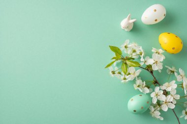 Pastel Paskalya sahnesinde beyaz bir tavşan, renkli bir yumurta ve nane arkaplanı karşısında çiçek açan çiçekler var. Paskalya kutlamaları ve mevsimlik kampanyalar için mükemmel bir görüntü.