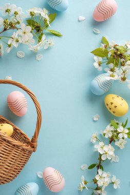 Pastel dikey Paskalya yumurtaları örülmüş bir sepetin içinde beyaz çiçeklerin ve taç yapraklarının arasında yumuşak mavi bir zemin üzerinde oturur ve Paskalya kutlamaları için şenlikli ve hoş bir sahne yaratır.