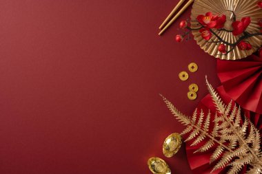 Zengin kırmızı arka planda altın sikkeler, dekoratif bir yelpaze, yemek çubukları ve Çin Yeni Yıl kutlamaları ve şenlikli selamlaşmaları çağrıştıran metalik aksanlar yer alıyor.