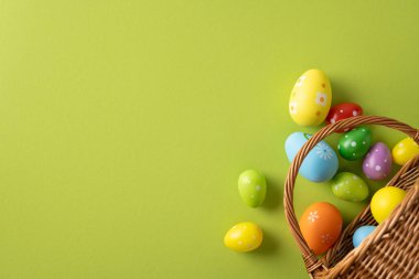 Renkli Paskalya yumurtaları parlak yeşil bir arka planda dokuma sepetinden dökülüyor. İlkbahar neşesini ve Paskalya kutlamaları ve tatil ruhunu çağrıştırıyor.