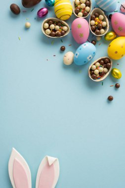 Mutlu Paskalya kutlamaları. Pastel süslemeli yumurtalar, çikolata kaplı kabuklar, serpiştirilmiş şekerlemeler ve bahar kutlaması için mavi arka planda düzenlenmiş neşeli tavşan kulakları.