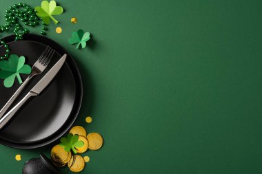 Bayram yemeği ve parti süslemeleri için yeşil arka planda siyah tabaklar, gümüş çatal bıçaklar, kağıt yoncalar ve altın paralarla Aziz Patrick Günü için bir masa düzenlemesi.