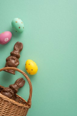 Çikolatalı tavşan figürleri ve nane yeşili arka planda hasır bir sepete boyanmış yumurtaların yer aldığı mutlu Paskalya dikey selamlaşmaları bahar kutlaması, tatil kartı tasarımı ve promosyon için mükemmel.