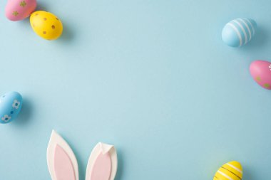 Pastel dekore edilmiş yumurtalar ve bahar kutlaması için yumuşak mavi arka planda düzenlenmiş neşeli tavşan kulaklarıyla mutlu Paskalyalar.