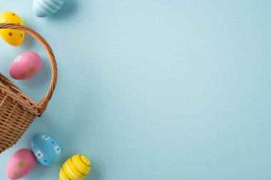 Hasır bir sepetten dökülen boyanmış yumurtalarla, bahar kutlamalarını ve renkli mevsimlik dekorları çağrıştıran yumuşak pastel arka planda mutlu Paskalya kutlamaları.
