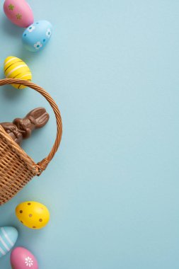 Mutlu Paskalya kutlaması ve yumuşak mavi bir arka planda pastel süslemeli yumurtayla hasır sepete yerleştirilmiş çikolatalı bir tavşanı selamlıyorum.