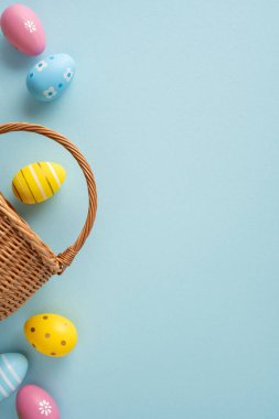 Soluk mavi arka planda hasır bir sepetten dökülen boyalı yumurtaların yer aldığı pastel bahar kutlaması için mutlu Paskalyalar.