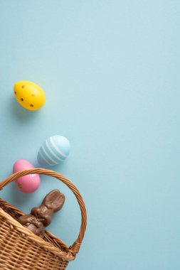 Mutlu Paskalya dikey selamlaması boyalı yumurtalar ve hasır sepete yerleştirilmiş çikolatalı tavşan yumuşak pastel arka planda kutlama, bahar sıcağı ve şenlikli dekorasyonu çağrıştırıyor.