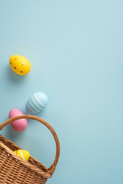 Mutlu Paskalya dikey kutlamaları. Pastel boyalı yumurtalar yumuşak mavi bir arka planda hasır sepetten dökülüyor. İlkbahar neşesini ve şenlikli mevsimsel dekorasyonu çağrıştırıyor.