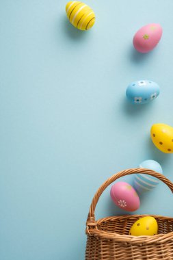 Hasır sepetten dökülen boyalı pastel yumurtalarla dolu mutlu Paskalya dikey selamlaşmaları bahar kutlamalarını ve şenlikli mevsimsel dekorları çağrıştıran yumuşak mavi bir fon üzerine.