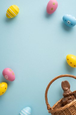 Dikey Mutlu Paskalya kutlamaları sahnesi pastel süslemeli yumurtalar hasır sepetin etrafına dizilmiş yumuşak mavi arka planda çikolatalı bir tavşan tutuyor.