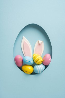 Yumurta şeklinde bir kağıt parçasına yerleştirilmiş tavşan kulakları ve rengarenk pastel yumurtalarla dolu mutlu Paskalya kutlamaları. Bahar kutlaması ve tatil dekorasyonu için.