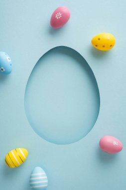 Pastel dikey dekore edilmiş yumurtalar, yumuşak mavi bir zemin üzerinde kağıttan bir yumurta çerçevesinin etrafını sarar ve kartlar, dekorasyonlar ve mevsimsel tasarımlar için Mutlu Paskalyalar ve bahar kutlamaları konseptini iletir.