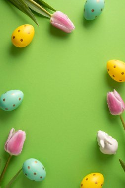 Pastel puantiyeli yumurta, pembe lale ve bahar kutlaması için parlak yeşil arka planda seramik tavşanın yer aldığı mutlu Paskalya dikey selamlama sahneniz kutlu olsun.