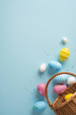 Süslü pastel yumurtaların yumuşak mavi bir arka planda hasır sepetten döküldüğü Paskalya 'nın dikey sahnesi bahar sıcağı ve şenlikli mevsimsel dekorasyonu çağrıştırıyor.