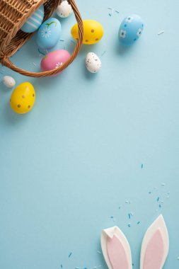 Hasır sepetten dökülen el boyalı yumurtalarla, köşeden bakan tavşan kulaklarıyla ve yumuşak mavi arka planda pastel konfetiyle mutlu Paskalya dikey sahneleri.