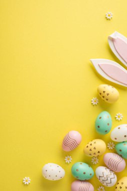 Pastel süslemeli yumurta, tavşan kulaklı dekor, papatya ve yumuşak sarı arka planlı bahar şenlik havasını taşıyan parlak Paskalya dikey sahnesi