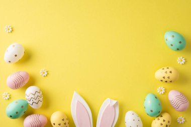 Happy Easter flat lay featuring pastel painted eggs, bunny ears and small daisies arranged on a bright yellow background for spring holiday inspiration