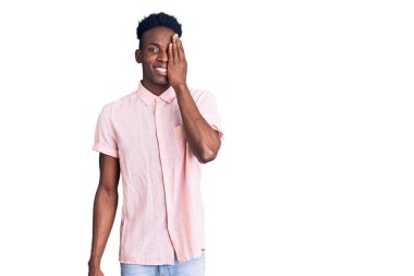 Genç Afro-Amerikan adam günlük kıyafetler giyiyor. Bir gözünü eliyle kapatıyor. Yüzünde kendine güvenen bir gülümseme ve sürpriz bir duygu var.. 