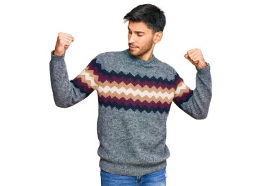 Sıradan bir kazak giyen genç yakışıklı adam, gururla gülümseyen kol kaslarını gösteriyor. fitness konsepti. 