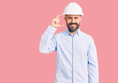 Mimar kasketi takan genç yakışıklı adam gülümsüyor ve kendine güvenen el hareketleriyle küçük boy işareti yapıyor parmaklarıyla bakıyor ve kameraya bakıyor. ölçüm kavramı. 