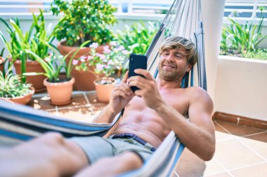 Orta yaşlı yakışıklı bir adam evinin terasında, bir hamakta telefonla dinleniyor.