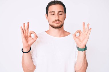 Genç yakışıklı adam, günlük beyaz tişört giyip rahatlıyor ve gözleri kapalı gülümsüyor parmaklarıyla meditasyon hareketi yapıyor. yoga konsepti. 
