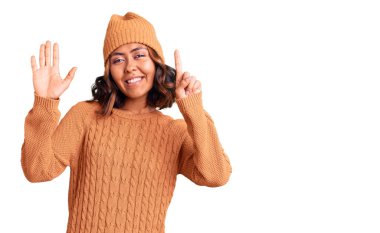 Genç, güzel, melez bir kadın yün kazak ve kış şapkası giyer ve altı numaralı parmaklarıyla işaret ederken kendinden emin ve mutlu bir şekilde gülümser.. 