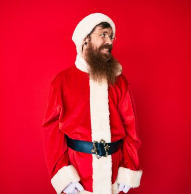 Uzun sakallı, yakışıklı, Noel Baba kostümü giyen, yüzünde gülümsemeyle yan yana bakan kızıl saçlı, doğal bir ifade. Kendinden emin gülen. 