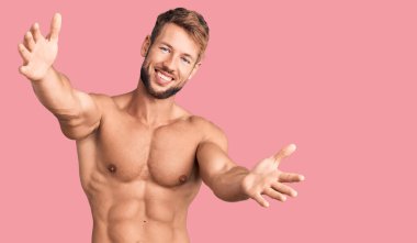 Beyaz bir genç, üstsüz bir şekilde kameraya bakıp sarılmak için kollarını açmış gülümsüyor. Mutluluğu kucaklayan neşeli bir ifade.. 