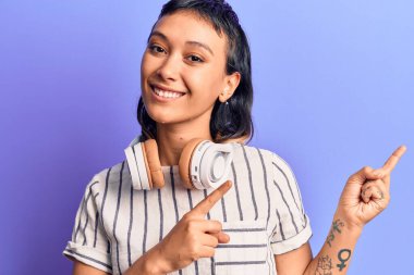 Kulaklıkla müzik dinleyen, gülümseyen ve kameraya bakan genç bir kadın iki eliyle ve parmaklarıyla kenarı işaret ediyor.. 