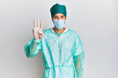 Cerrah üniforması giyen ve tıbbi maske takan genç yakışıklı adam üç numarayı gösterip işaret ederken kendinden emin ve mutlu gülümsüyor.. 