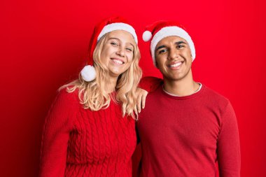 Irklar arası genç çift Noel şapkası takıyor. Olumlu ve mutlu görünüyorlar. Ayakta duruyorlar ve gülümsüyorlar. 
