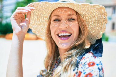 Yaz şapkası takmış, mutlu bir şekilde gülümseyen güzel sarışın bir kadın. Şehrin caddesinde yüzünde gülümsemeyle dikilirken.