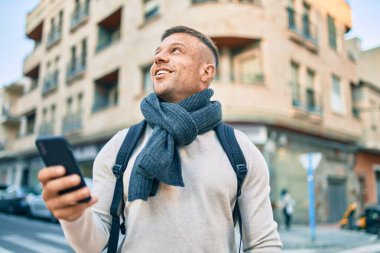Genç beyaz bir öğrenci şehirde akıllı telefon kullanarak mutlu bir şekilde gülümsüyor..