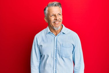 Orta yaşlı, gri saçlı, günlük kıyafetler giyen, yüzünde gülümsemeyle yan yana bakan, doğal bir ifade. Kendinden emin gülen. 