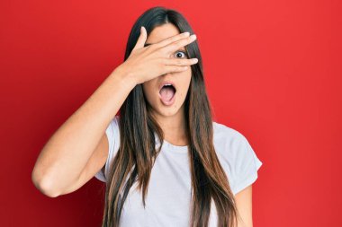 Genç esmer kadın, kırmızı arka planda günlük beyaz tişört giyiyordu. Şok olmuş bir şekilde yüzünü ve gözlerini kapatıyordu. Utanmış bir ifadeyle parmaklarının arasından bakıyordu.. 