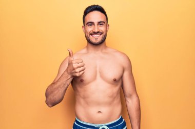 Sakallı, kolsuz tişörtlü, sarı arka planda duran mutlu ve pozitif gülümseyen, başparmağını kaldırmış, mükemmel ve onay işareti yapan genç yakışıklı bir adam.