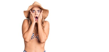 Bikini giyen, şapkalı, korkmuş, şok olmuş, şaşırmış ve şaşırmış genç, sarışın bir kadın. 