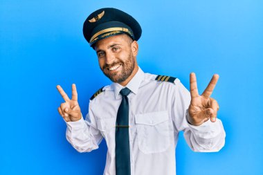 Sakallı, uçak pilotu üniforması giyen, gülümseyen ve parmaklarını zafer işareti yaparken gösteren yakışıklı bir adam. İki numara.. 
