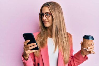 Genç sarışın kadın akıllı telefon kullanıyor ve bir fincan kahve içiyor. Gülümsüyor, yan tarafa bakıyor ve düşünüyor.. 