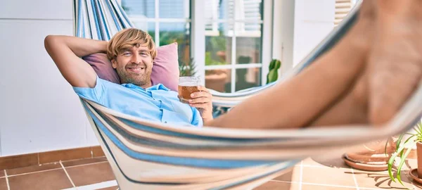 中年英俊男子躺在自家的阳台上 悠闲地躺在吊床上 喝着新鲜啤酒 — 图库照片